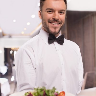 Cheerful waiter. Handsome young waiter in shirt and bow tie holding a plate with salad and smiling